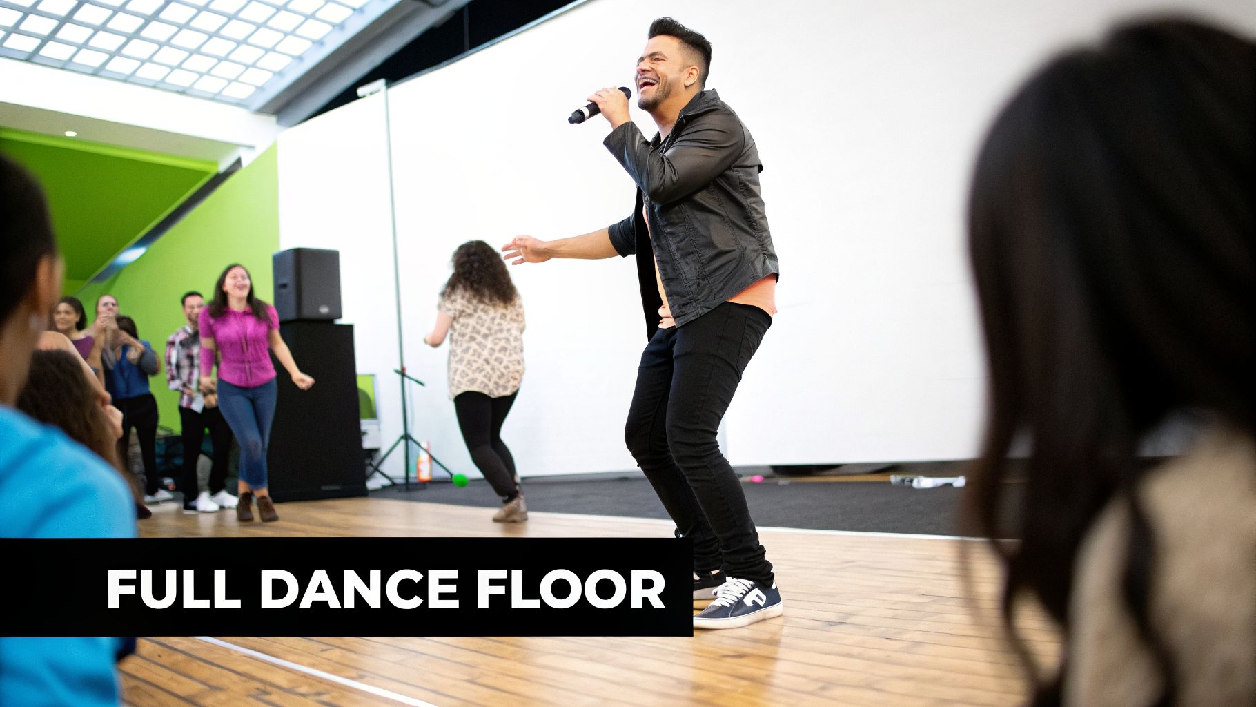 A smiling man sings into a microphone while dancing on a wooden floor, surrounded by a joyful crowd.