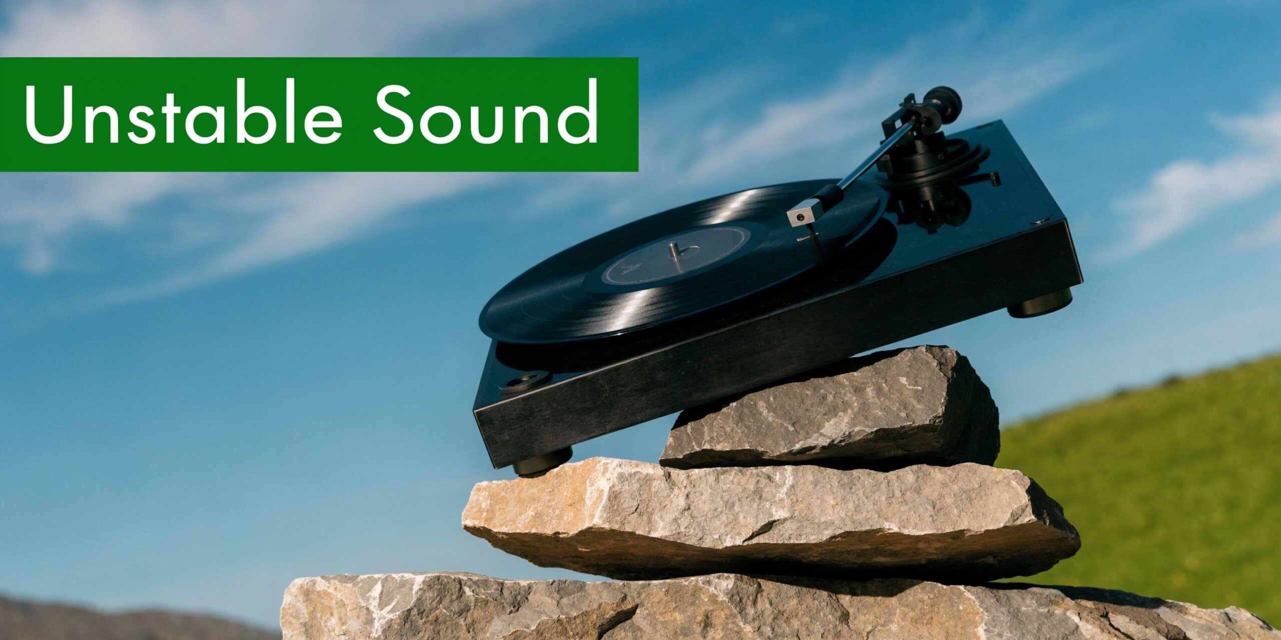 A black record player balanced precariously on a stack of flat stones against a blue sky.